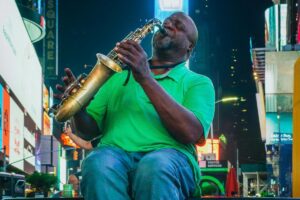 A man in a green shirt plays a saxophone with passion in the vibrant setting of a city square, surrounded by bright billboards. Similar to how his music draws people in, BaoLiba aims to connect Australian influencers and brands to the world, creating a harmonious blend and drawing global attention.
