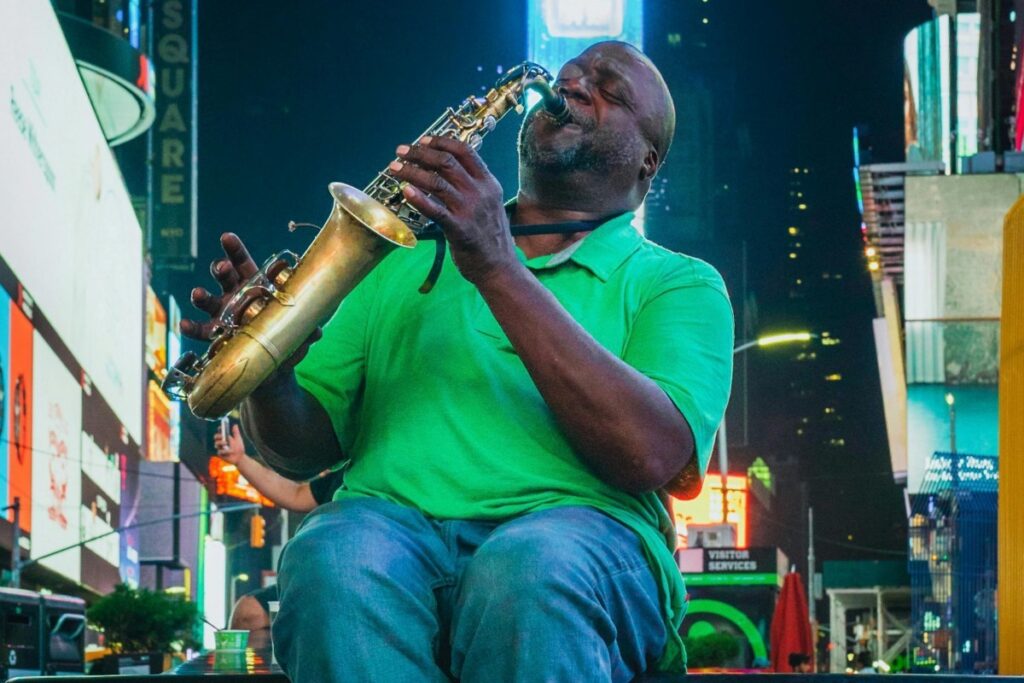 A man in a green shirt plays a saxophone with passion in the vibrant setting of a city square, surrounded by bright billboards. Similar to how his music draws people in, BaoLiba aims to connect Australian influencers and brands to the world, creating a harmonious blend and drawing global attention.