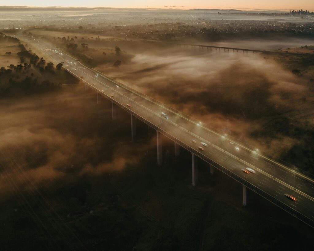 An aerial view of a long bridge stretching across a landscape shrouded in mist at dawn in Australia, with vehicles moving along it. Just as this bridge connects different areas, "Reddit Australia: Engaging Local Communities" serves as a link to connect and interact with various local groups on the Reddit platform in Australia.
