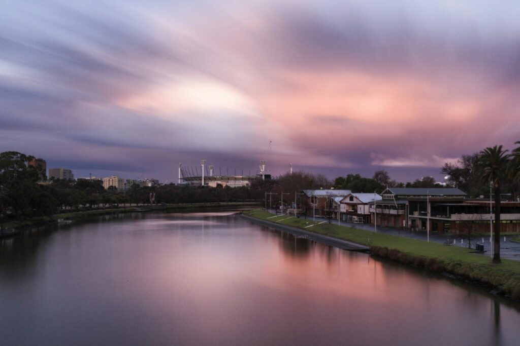 A serene view of a river in Australia, with buildings and palm trees lining its banks. In the distance, a stadium looms under a sky painted with hues of purple and pink. This peaceful scene is a great backdrop to learn about "The Ultimate Facebook Ads Guide for Australian Businesses", which can help local enterprises reach their target audience effectively.