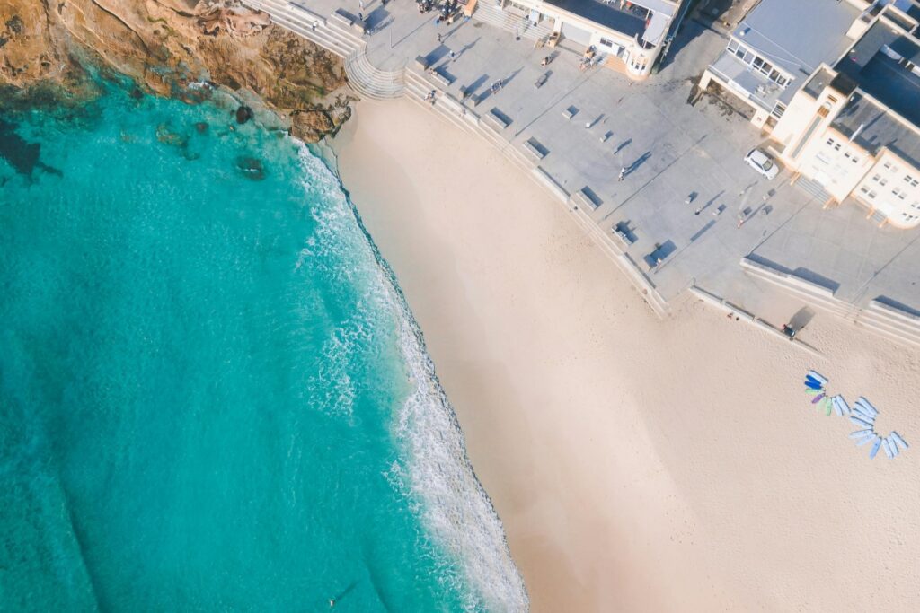 This aerial image showcases a beautiful Australian beach with crystal - clear turquoise waters gently lapping against a sandy shore. Alongside the beach, there are modern buildings and a paved promenade where a few people can be seen. Some surfboards are neatly arranged on the sand. For those aiming for "YouTube Success in Australia Tips for Creators", if your content is about travel, beach sports, or Australian lifestyle, this kind of image can be great for thumbnails. You can create videos about beach activities like surfing lessons, exploring the coastal architecture, or sharing the best times to visit such beaches. Use relevant keywords and hashtags like #AustralianBeaches, #SurfingInAustralia, and #CoastalLifeAustralia in your video titles, descriptions, and tags. Engage with the local Australian YouTube community, collaborate with other creators, and regularly upload high - quality content to increase your chances of success on the platform.