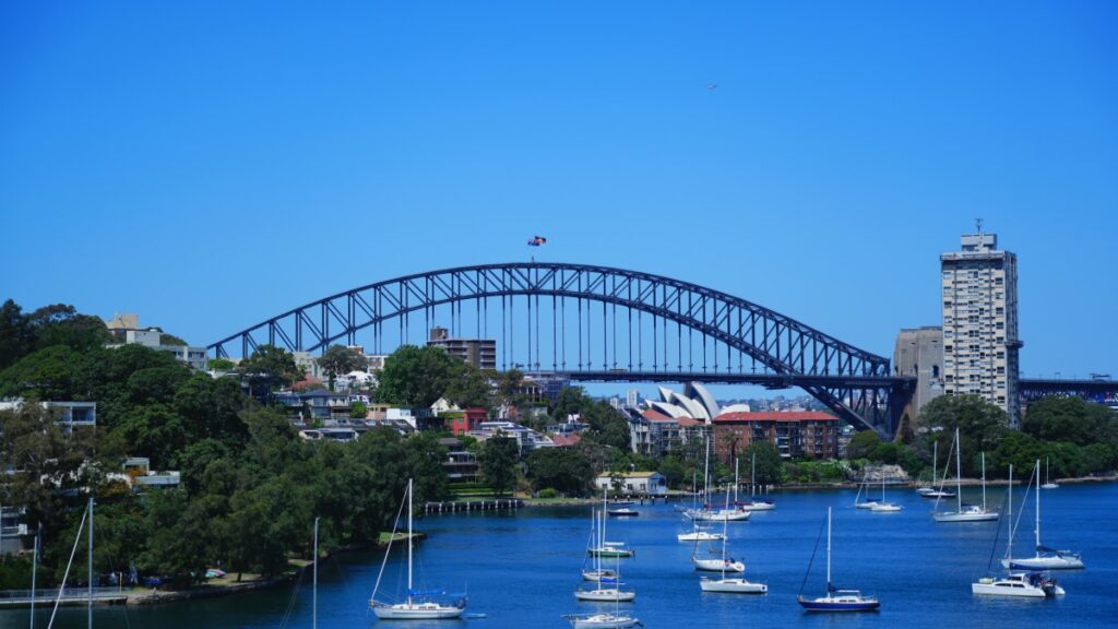This picturesque scene showcases the iconic Sydney Harbour Bridge in Australia, with its majestic arch standing tall against a clear blue sky. Below, the calm waters are dotted with sailboats, surrounded by lush greenery and charming buildings. For those interested in making the most of their time Down Under, check out "How to Earn Money on Bigo Live A Comprehensive Guide" to explore ways to monetize your passions while enjoying Australia's beauty.